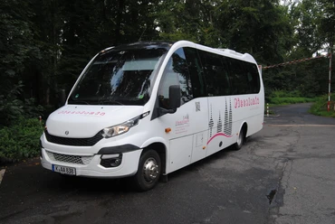 Bergischer WanderBus Ein weißer Reisebus mit Logos, vor Bäumen auf einer asphaltierten Straße in ruhiger Umgebung geparkt.