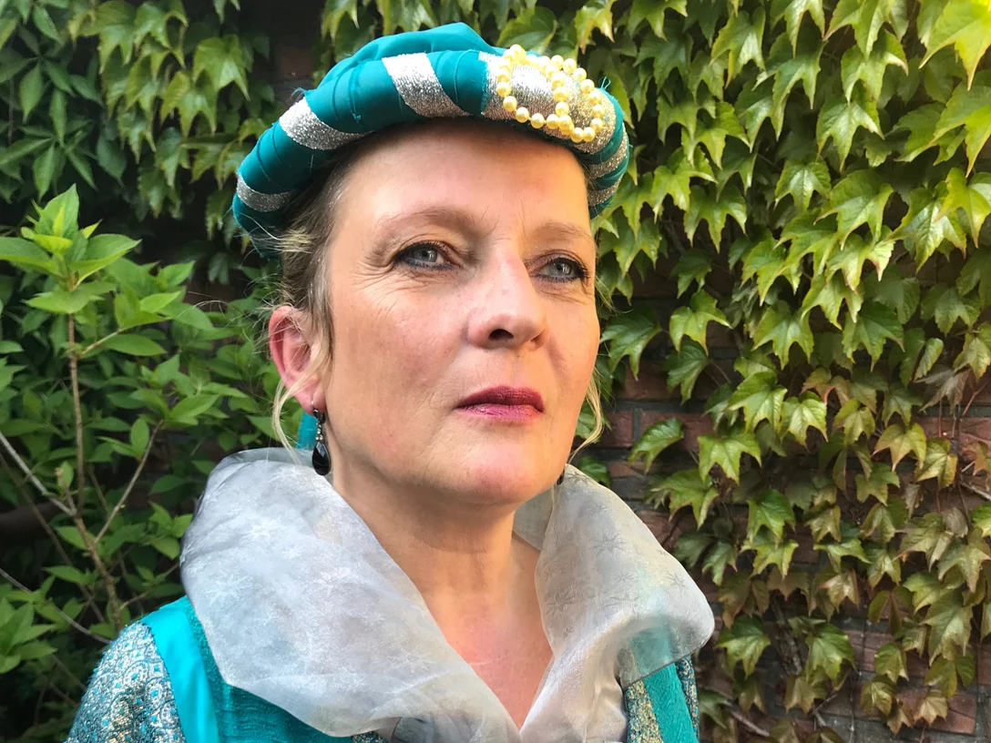 LAK: Der letzte Häuptling - Der Mann von Gräfin Theda Frau in historischem Kostüm mit türkis-silberner Haube und besticktem Kleid vor grüner Efeuwand.