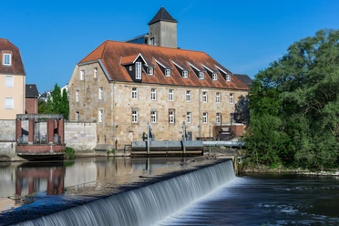 Rheine_Emswehr 2_Foto_ Rheine Tourismus Veranstaltungen eV_Y Mokdad.jpg Historisches Gebäude mit rotem Ziegeldach und Emswehr im Vordergrund bei sonnigem Wetter in Rheine.