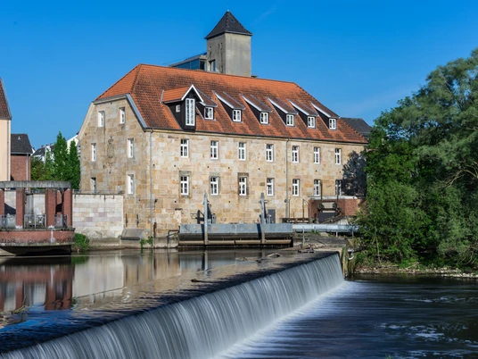 Rheine_Emswehr 2_Foto_ Rheine Tourismus Veranstaltungen eV_Y Mokdad.jpg Historisches Gebäude mit rotem Ziegeldach und Emswehr im Vordergrund bei sonnigem Wetter in Rheine.