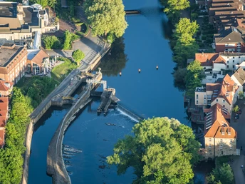 Blick von oben auf das Emswehr in Rheine, umgeben von üppigem Grün und Gebäuden links und rechts.