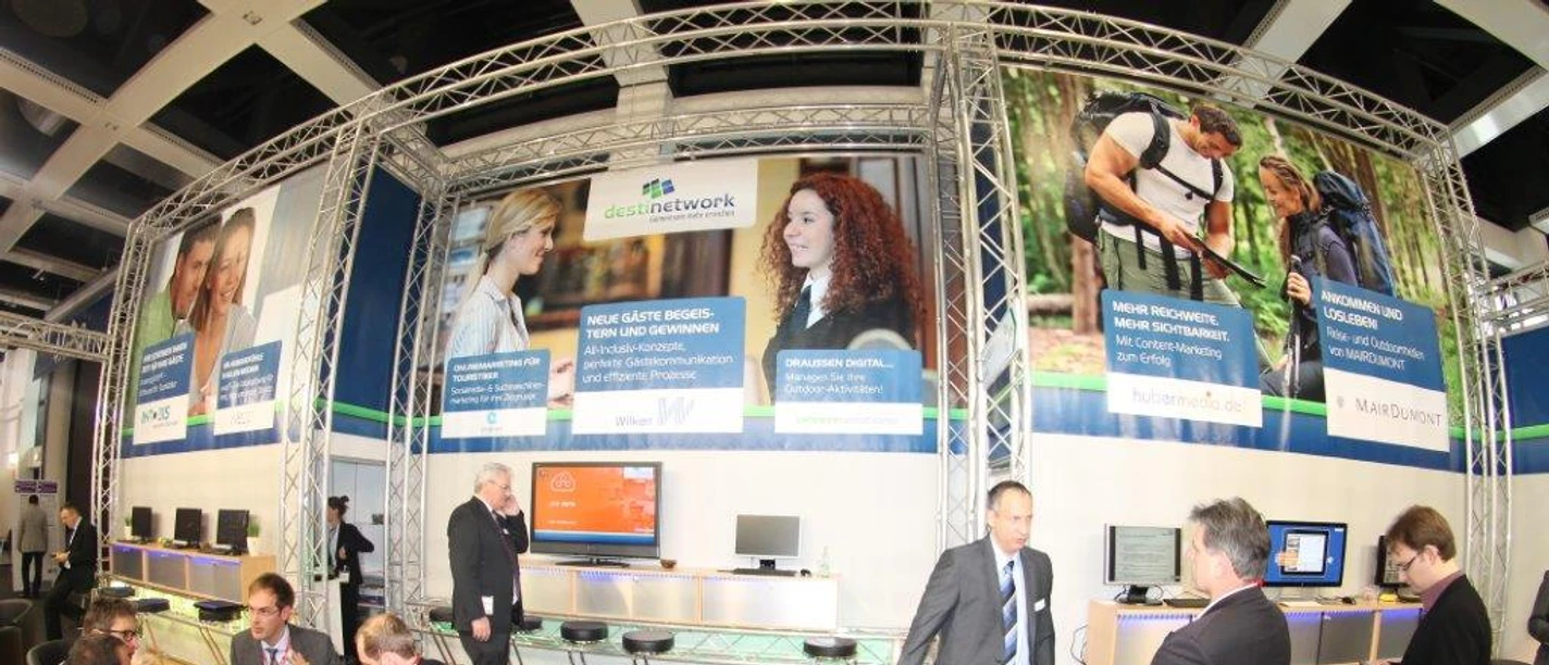 testbild Herzlich Willkommen auf einer Messe. Die Szene zeigt Menschen im Gespräch an einem Informationsstand.