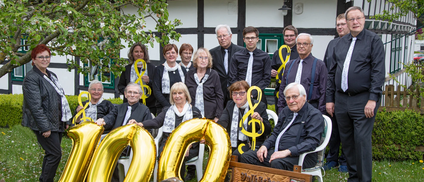 Mitglieder des Volkschors Gehlenbeck feiern vor einem Fachwerkhaus ein Jubiläum mit goldenen Luftballons.