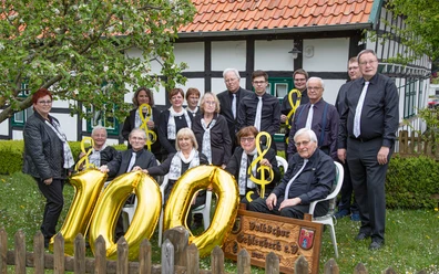 VolkschorGehlenbeck.JPG Mitglieder des Volkschors Gehlenbeck feiern vor einem Fachwerkhaus ein Jubiläum mit goldenen Luftballons.
