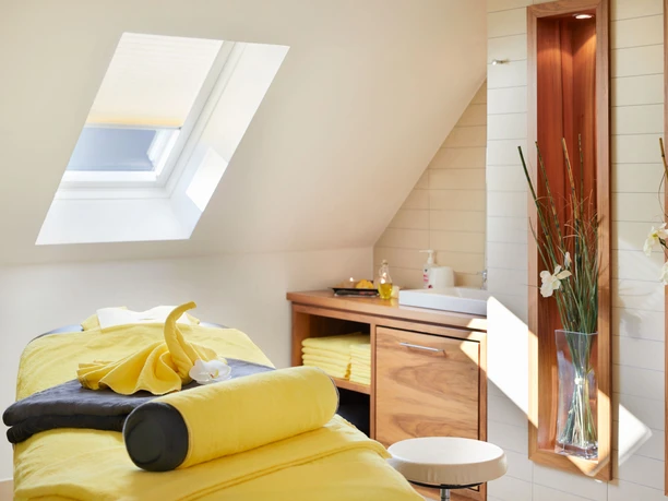 Bright massage room with yellow lounger, towels and orchid, skylight, wooden cabinet with washbasin, relaxed atmosphere.