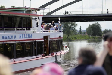 Das Fahrgastschiff Helena auf der Weser, im Hintergrund eine Brücke und grüne Uferbereiche.