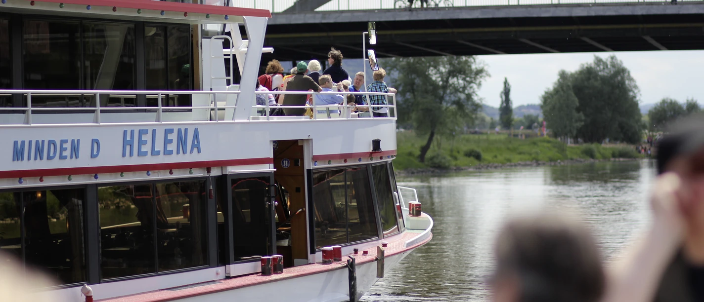 Das Fahrgastschiff Helena auf der Weser, im Hintergrund eine Brücke und grüne Uferbereiche.
