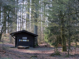 Herlingsburg, GN.JPG Eine Holzhütte steht umgeben von hohen, lichten Bäumen im Wald, mit einem Baumstamm als Sitzplatz.