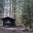 Herlingsburg, GN.JPG Eine Holzhütte steht umgeben von hohen, lichten Bäumen im Wald, mit einem Baumstamm als Sitzplatz.