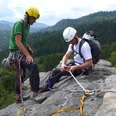 Klettertour Ottendorfer Hütte Zwei Kletterer mit Helmen und Ausrüstung stehen auf einem Felsen vor einer bewaldeten Berglandschaft; einer kniet und sichert ein Seil.