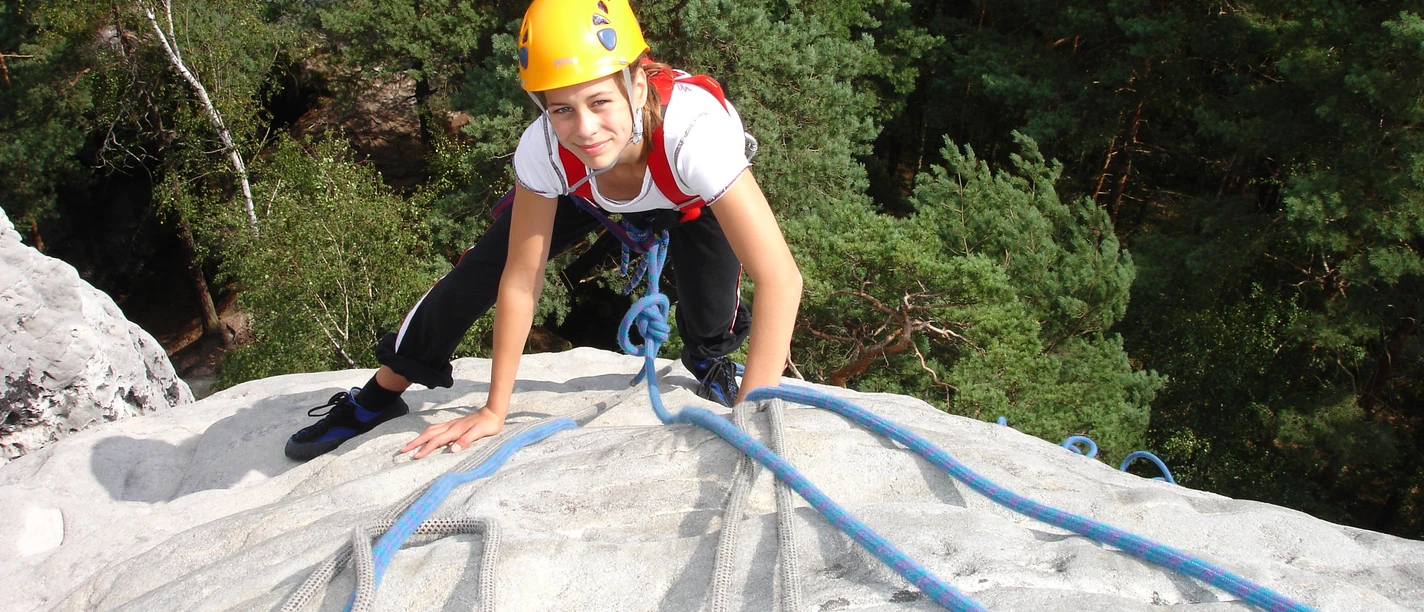 Kletterschule Lilienstein Eine Person mit gelbem Helm klettert auf einem Felsen, umgeben von grünen Bäumen, und hält sich an blauen Seilen fest.