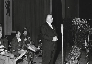 Kurdirektor Hans-Otto-Schmidt Hans-Otto-Schmidt steht in einem feierlichen Saal am Mikrofon, umgeben von Musikern und Blumenschmuck.