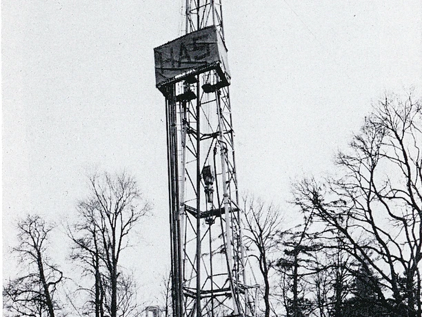Alexander-von-Humboldt-Sprudel Ein hoher, metallischer Turm zur Erdölerschließung, umgeben von kahlen Bäumen und bewölktem Himmel.