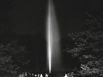 Nachts leuchtender Wassersprudel in Bad Oeynhausen, umgeben von Bäumen und Weglichtern.