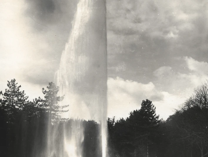 Hoher Wasserstrahl des Jordan-Sprudels, von Bäumen umgeben, mit zwei Personen im Vordergrund.