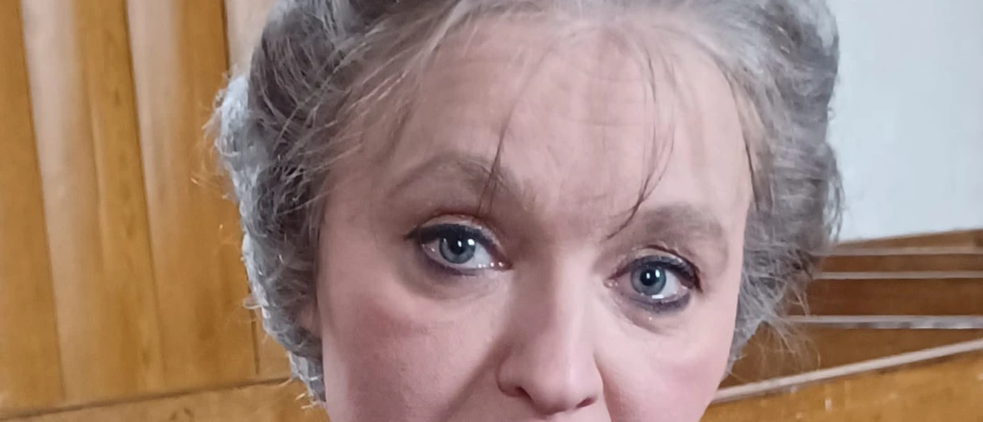 Dörfer erzählen Geschichten: Wilhelmine Siefkes Frau in historischer Kleidung mit grauer Hochsteckfrisur in einem Holzsaal, Porträtaufnahme.