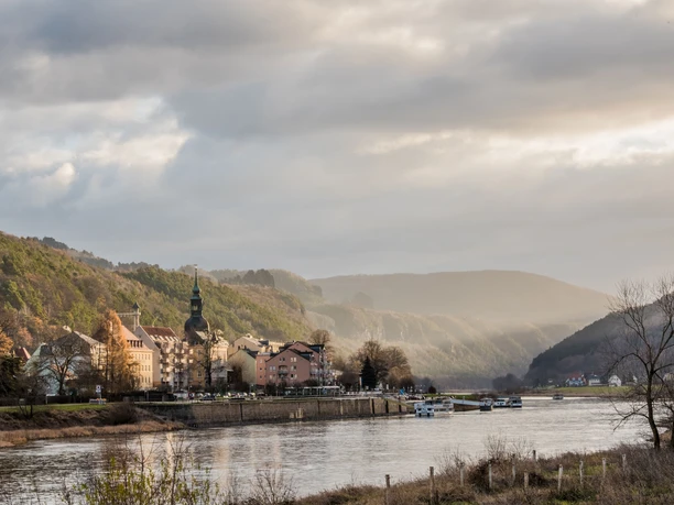 Small riverside town with historic buildings and church tower, surrounded by wooded hills under a cloudy sky.