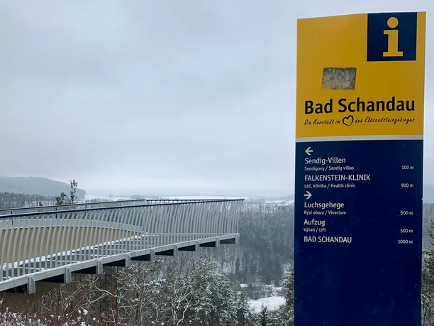 Geel informatiebord met het opschrift "Bad Schandau" voor een besneeuwd landschap en een moderne brug op de achtergrond.