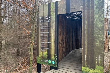 Holzsteg führt durch einen überdachten Weg im Wald, mit einem Schild "Weg zur Wildnis" in mehreren Sprachen, umgeben von kahlen Bäumen.