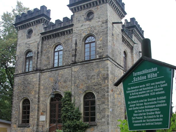 Stone tower castle with battlements and arched windows, next to it a green sign with information about the building, surrounded by trees.