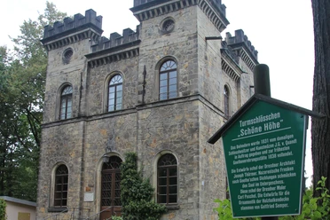 Belvedere Schöne Höhe Steinernes Turmschlösschen mit Zinnen und Rundbogenfenstern, daneben ein grünes Schild mit Informationen über das Gebäude, umgeben von Bäumen.