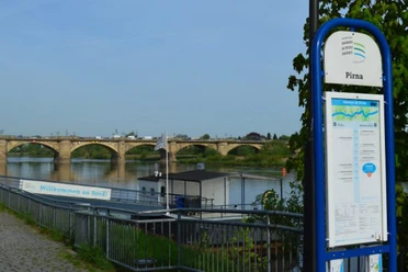 Gezicht op een stenen brug en de aanlegsteiger van de Saxon Steamship Company over een rivier in Pirna, met een informatiebord op de voorgrond en een heldere hemel.