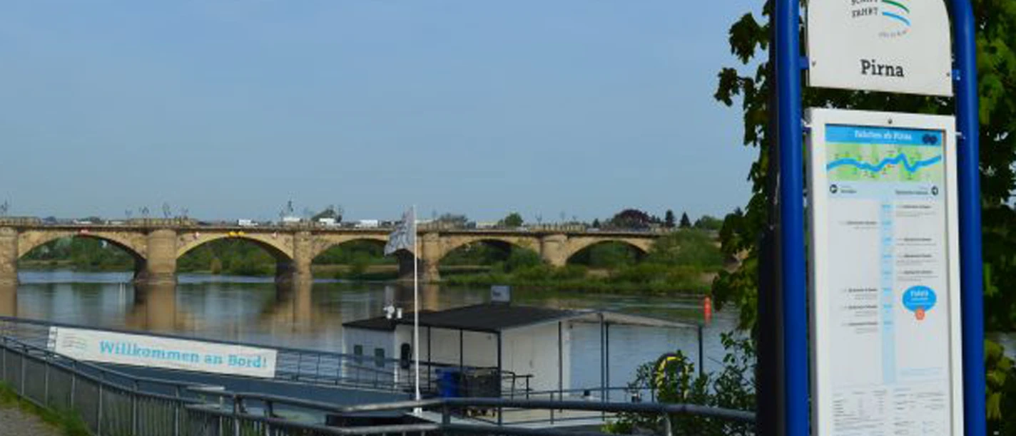 View of a stone bridge and the jetty of the Saxon steamboat company across a river in Pirna, with an information sign in the foreground and a clear sky.