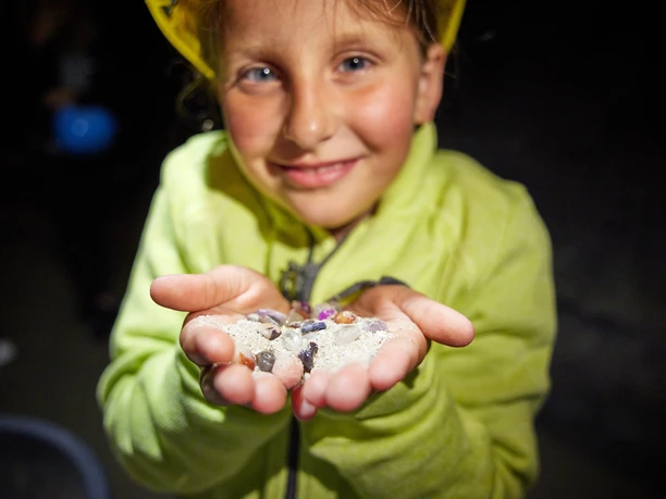 Edelsteine finden bei einer Kinderführung Ein lächelndes Kind in leuchtend gelber Jacke präsentiert stolz glitzernde Edelsteine in seinen Händen.