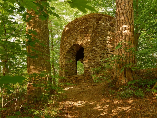 Goßdorfer Raubschloss Steinruine mit rundem Dach im Wald, umgeben von hohen Bäumen und grünem Laub, im warmen Sonnenlicht.