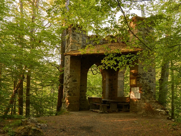 Goßdorf Robbery Castle Stone ruins surrounded by green trees, with a wooden table and benches underneath. The atmosphere is quiet and idyllic.