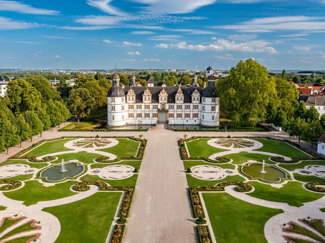 Paderborn-SchlossNeuhaus-Teutoburger-Wald-Tourismus-Patrick-Gawandtka-125-CC-BY-SA.jpg Luftaufnahme des Schlosses Neuhaus in Paderborn, umgeben von symmetrischen Gärten und Grünflächen.