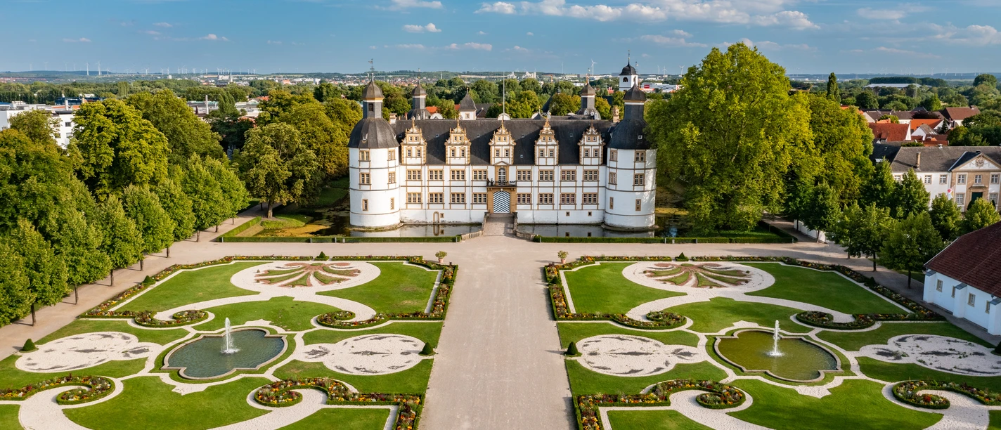 Paderborn-SchlossNeuhaus-Teutoburger-Wald-Tourismus-Patrick-Gawandtka-125-CC-BY-SA.jpg Luftaufnahme des Schlosses Neuhaus in Paderborn, umgeben von symmetrischen Gärten und Grünflächen.