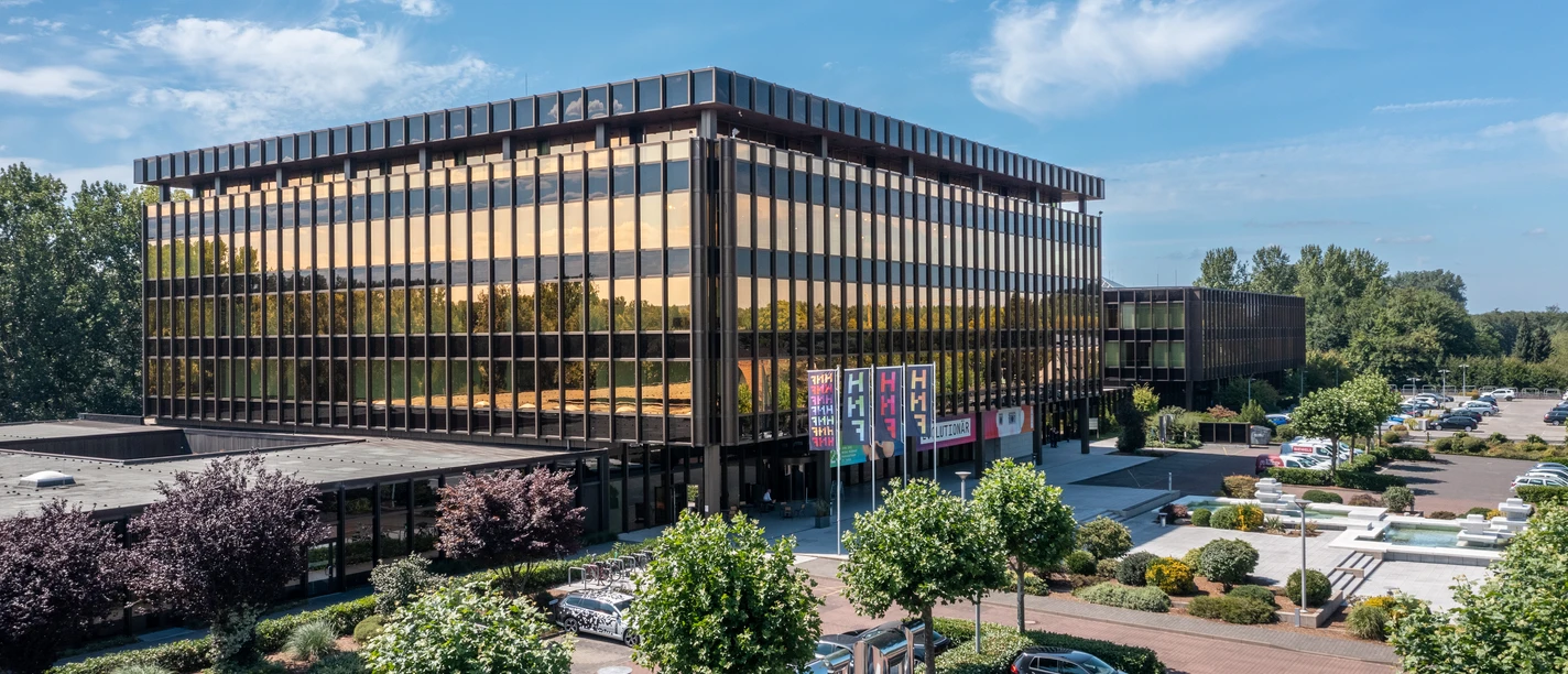 Paderborn-NixdorfMuseum-Teutoburger-Wald-Tourismus-Patrick-Gawandtka-082-CC-BY-SA.jpg Modernes, gläsernes Bürogebäude in Paderborn, umgeben von Bäumen und geparkten Autos bei klarem Himmel.