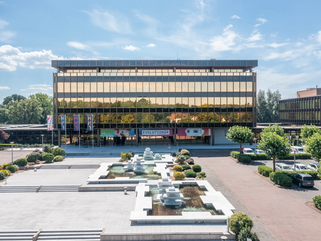 Paderborn-NixdorfMuseum-Teutoburger-Wald-Tourismus-Patrick-Gawandtka-084-CC-BY-SA.jpg Gebäude mit gläserner Fassade, davor Brunnenanlage und bepflanzter Vorplatz bei sonnigem Wetter.