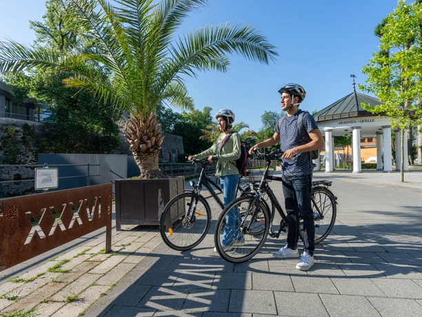 BadLippspringe-Lippequelle-Teutoburger-Wald-Tourismus-Patrick-Gawandtka-014.jpg Zwei Radfahrer stehen vor einer Palmenpflanze und der Arminius-Quelle in Bad Lippspringe.