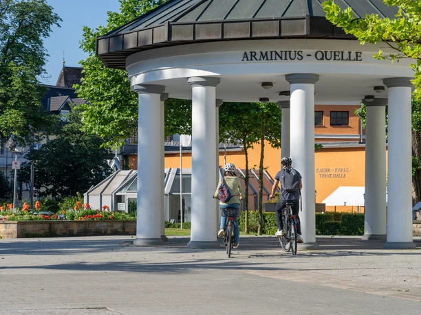 <p>Zwei Radfahrer fahren unter dem Pavillon der Arminiusquelle in Bad Lippspringe bei Sonnenschein.</p>