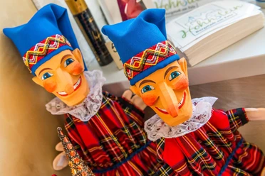 Hohnsteiner Kasper Zwei bunte Kasperlefiguren mit blauen Mützen und karierten Kleidern stehen nebeneinander, fröhlich lächelnd, auf einem Regal.