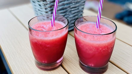 Zwei rote Smoothies mit gestreiften Strohhalmen auf einem Holztisch, im Hintergrund ein Korb.