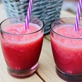 Zwei rote Smoothies mit gestreiften Strohhalmen auf einem Holztisch, im Hintergrund ein Korb.
