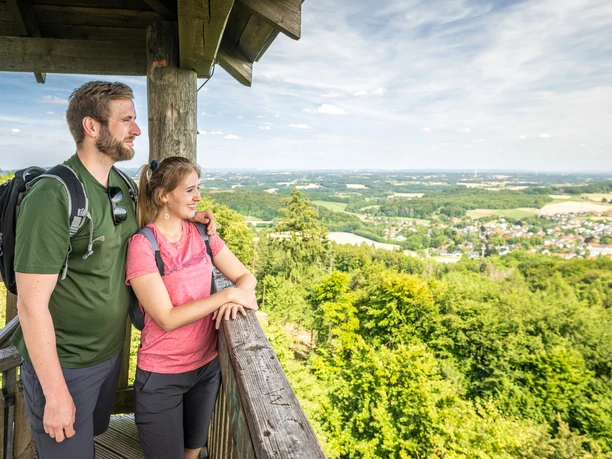 Ein Paar steht auf einem Aussichtsturm, genießt weite Ausblicke über grüne Wälder und Landschaften.