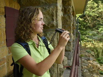 Steinhagen-Schwedenschanze-Teutoburger-Wald-Tourismus-F-Grawe (92).JPG Eine junge Frau mit Fernglas steht an einem Steinhaus, blickt lächelnd in die dichte Waldlandschaft.