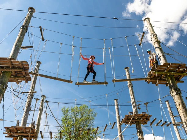 Bielefeld-Kletterpark-Teutoburger-Wald-Tourismus-D-Ketz-026.jpg Ein Hochseilgarten mit blauen Himmel, ein Abenteuerlustiger überquert eine Seilbrücke in luftiger Höhe.