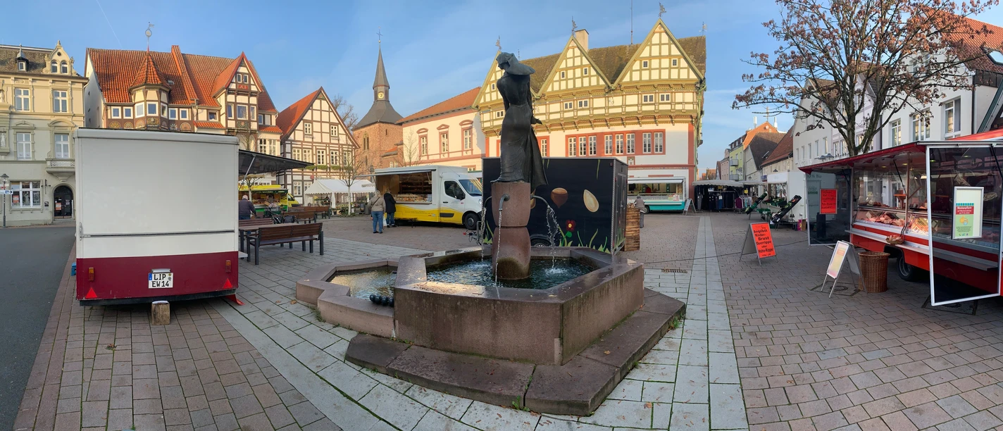 Marktplatz mit Brunnen, umgeben von historischen Fachwerkhäusern und Marktständen bei sonnigem Wetter.