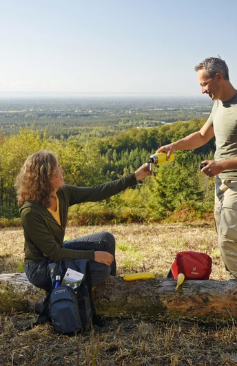 Zwei Personen auf einer Lichtung im Teutoburger Wald, tauschen Getränke mit bewaldetem Lipperland im Hintergrund.