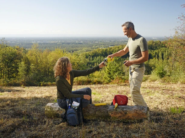 Oerlinghausen-Ausblick-ins-Lipperland-Teutoburger-Wald-Tourismus-F-Grawe (64).JPG Zwei Personen auf einer Lichtung im Teutoburger Wald, tauschen Getränke mit bewaldetem Lipperland im Hintergrund.