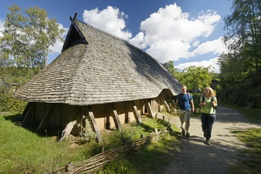 Rekonstruiertes Langhaus aus Holz mit Strohdach, zwei Personen spazieren auf einem Waldweg vorbei.