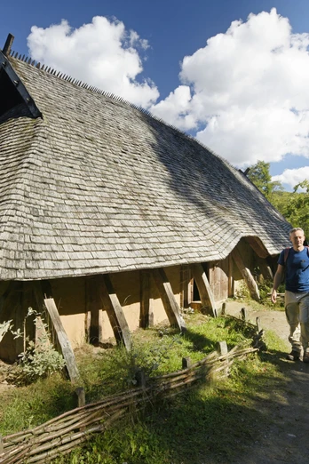 Rekonstruiertes Langhaus aus Holz mit Strohdach, zwei Personen spazieren auf einem Waldweg vorbei.