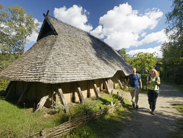 Rekonstruiertes Langhaus aus Holz mit Strohdach, zwei Personen spazieren auf einem Waldweg vorbei.