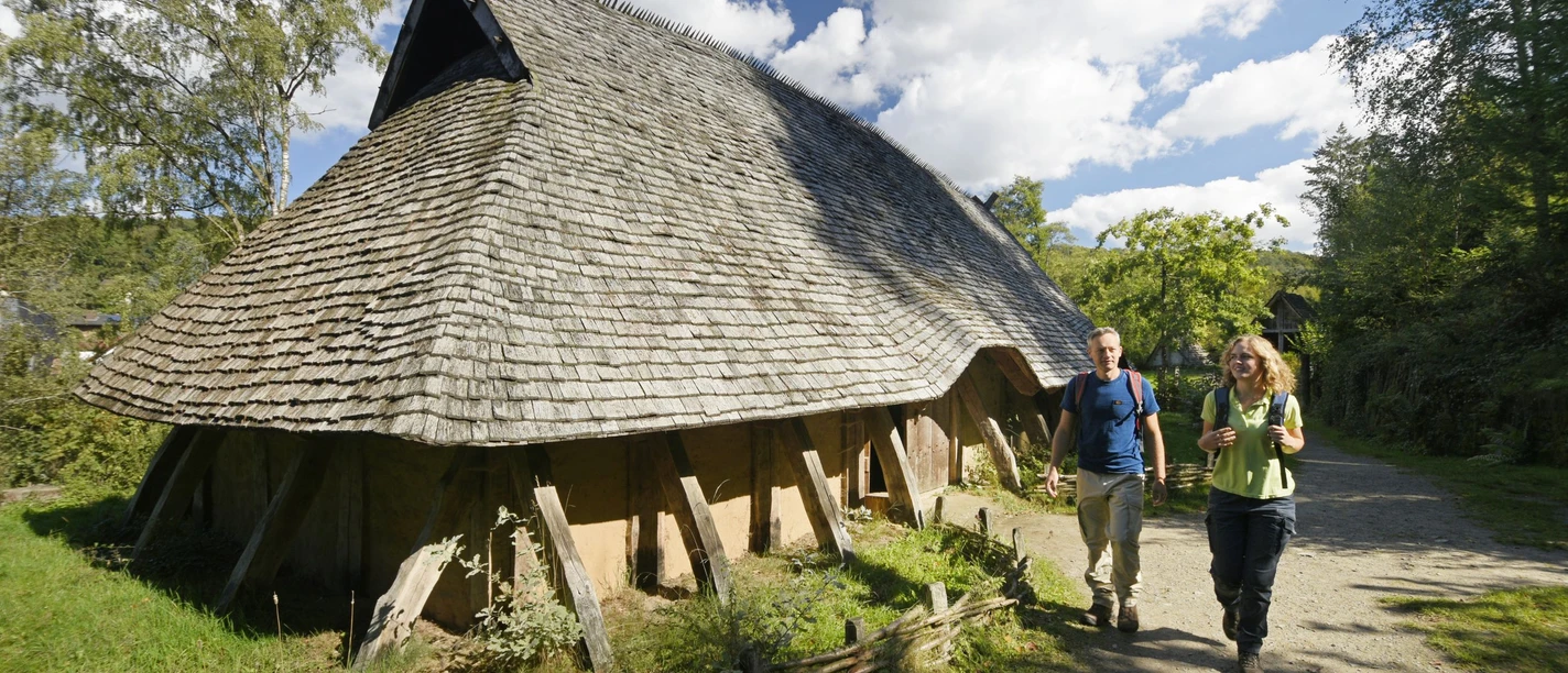 Oerlinghausen-AFM-Langhaus-Teutoburger-Wald-Tourismus-F-Grawe (50)_klein.jpg Rekonstruiertes Langhaus aus Holz mit Strohdach, zwei Personen spazieren auf einem Waldweg vorbei.