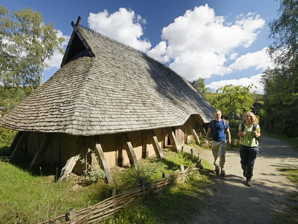 Oerlinghausen-AFM-Langhaus-Teutoburger-Wald-Tourismus-F-Grawe (50)_klein.jpg Rekonstruiertes Langhaus aus Holz mit Strohdach, zwei Personen spazieren auf einem Waldweg vorbei.